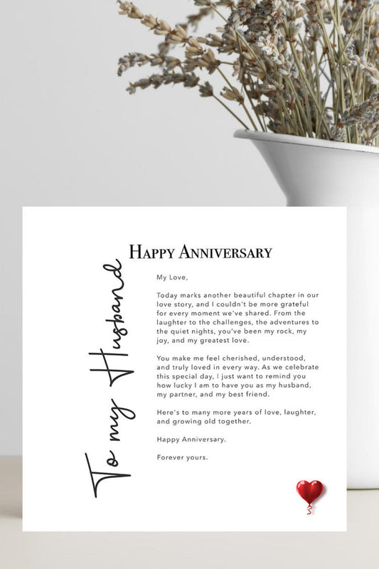 A printed anniversary card with a sentimental message addressed 'To my Husband' and 'Happy Anniversary,' styled in a flat-lay setting next to a white vase with dried flowers on a light gray surface.