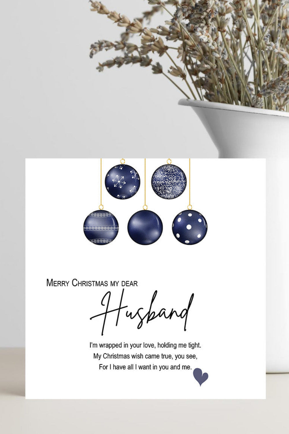A white Christmas greeting card with navy blue hanging ornaments and a poem addressed to a husband, styled on a light surface beside a white vase with dried flowers.