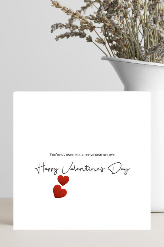 A square white greeting card with black script text reading 'Happy Valentine's Day' and two small red hearts, styled in front of a white vase with dried flowers on a light surface.
