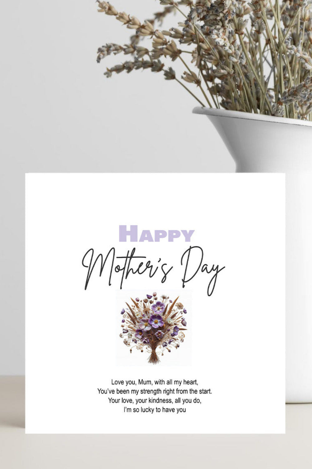 A square Mother's Day greeting card with a white background, featuring the text 'Happy Mother's Day' in lavender and black script, a small purple floral tree illustration, and a short poem below. The card is styled in a home setting on a light surface next to a white vase containing dried flowers.