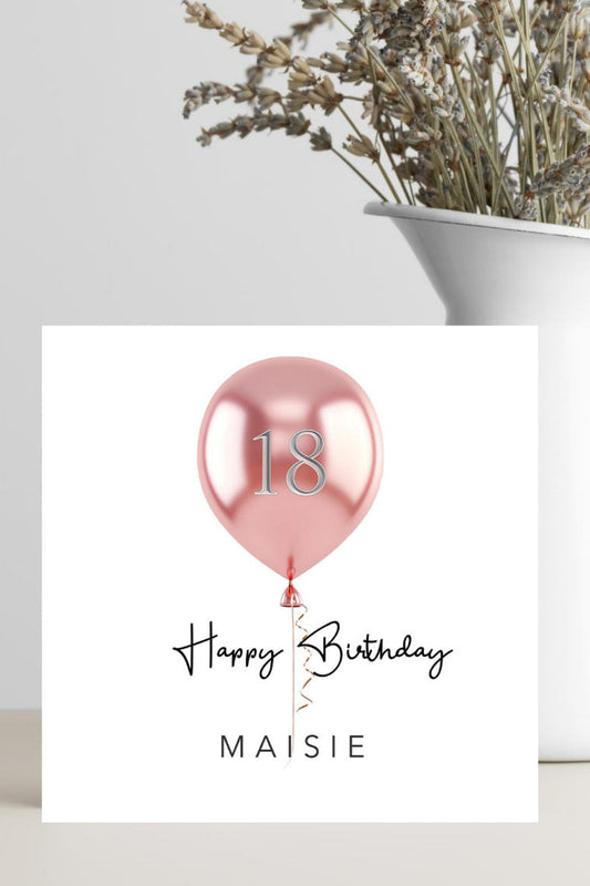A square birthday card with a metallic pink balloon graphic and the text '18 Happy Birthday MAISIE' is displayed on a tabletop next to a white vase with dried flowers.
