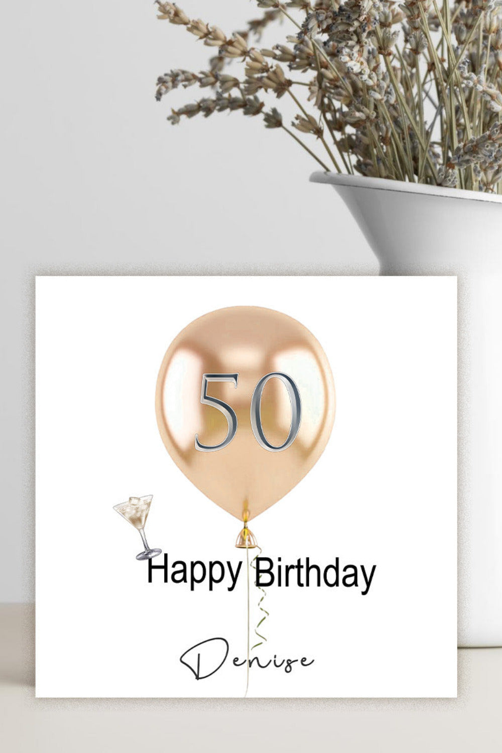 A square white birthday card with a metallic rose-gold balloon graphic containing the number 50, plus the text 'Happy Birthday' and the name 'Denise', displayed on a light surface next to a white vase with dried flowers.