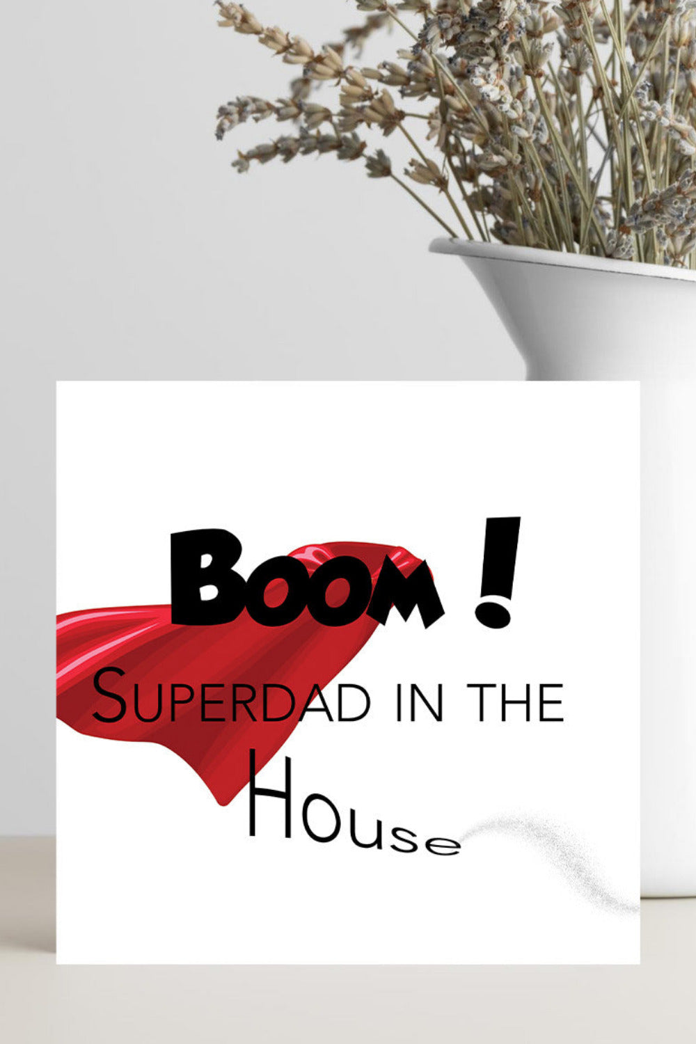 A square white card with the text 'BOOM! SUPERDAD IN THE House' in black, overlaid with a red superhero cape graphic, standing on a light surface beside a white vase with dried flowers.