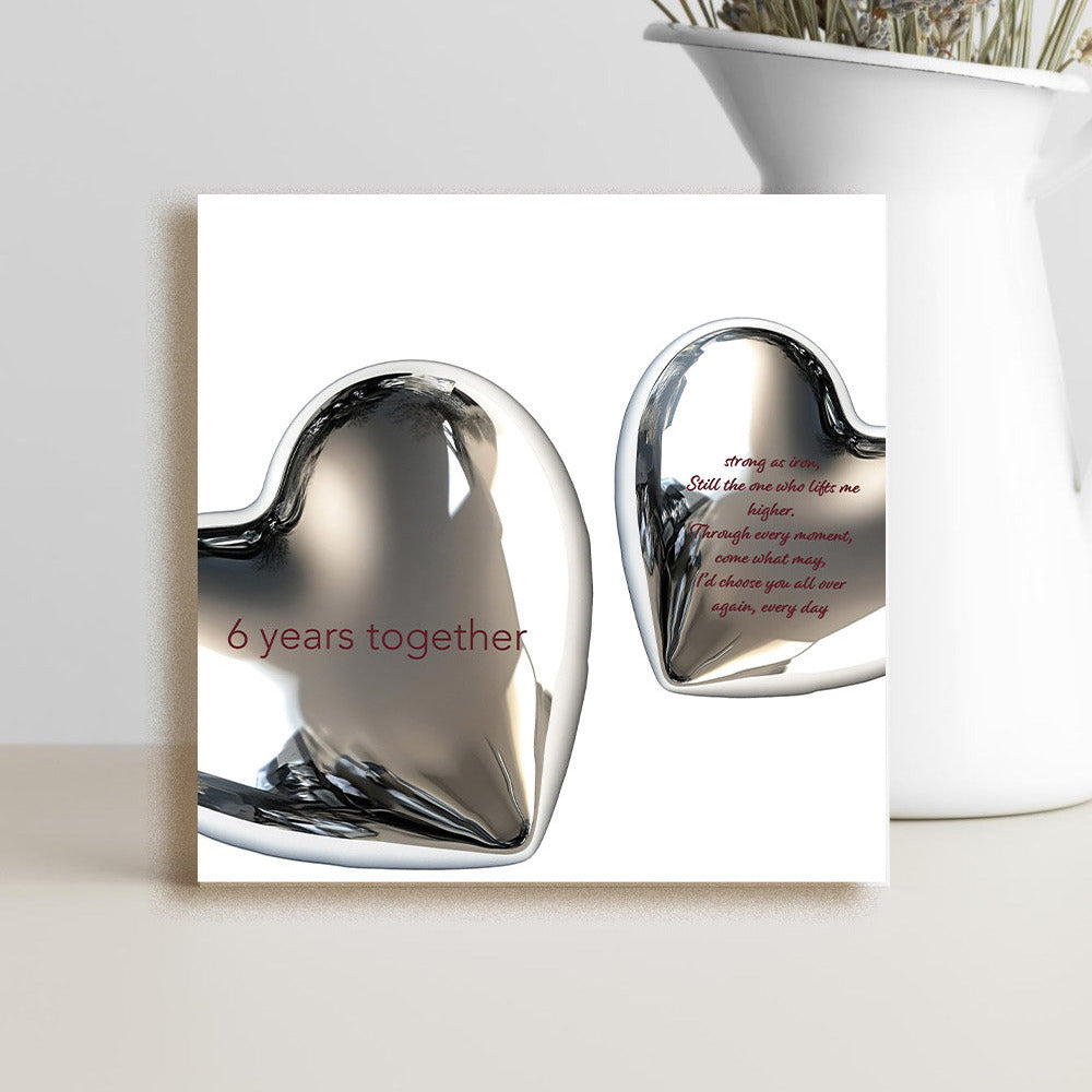 A square greeting card or print featuring two reflective metallic hearts on a white background, with one heart reading '6 years together' in red. The card is styled on a light surface beside a white vase with dried flowers against a light gray wall.