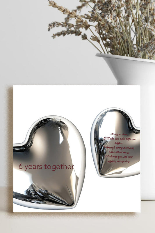 A square greeting card or print featuring two reflective metallic hearts on a white background, with one heart reading '6 years together' in red. The card is styled on a light surface beside a white vase with dried flowers against a light gray wall.