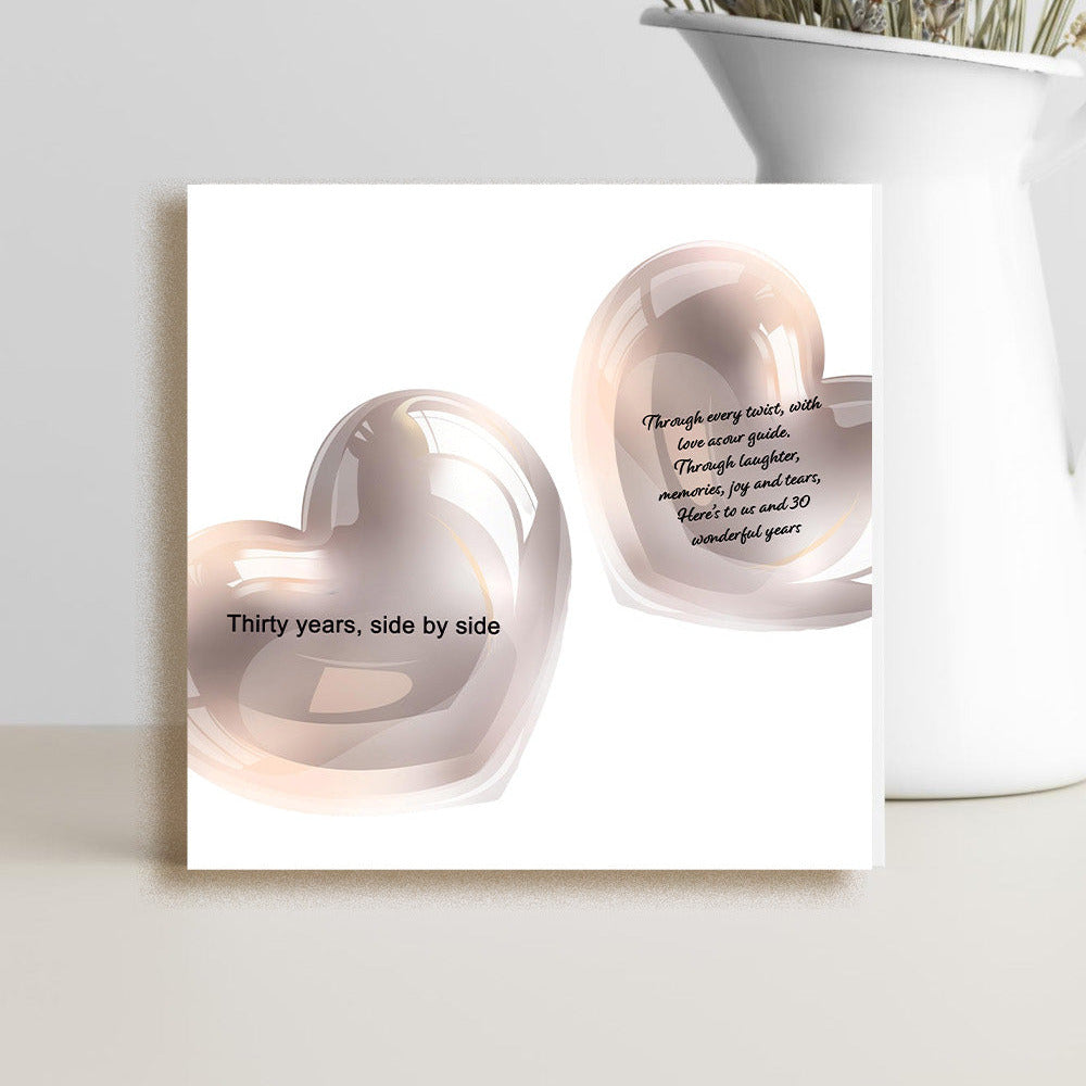 A square white art print or card featuring two glossy blush pink heart shapes with black text, displayed on a light surface beside a white vase holding dried flowers.