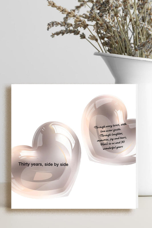 A square white art print or card featuring two glossy blush pink heart shapes with black text, displayed on a light surface beside a white vase holding dried flowers.