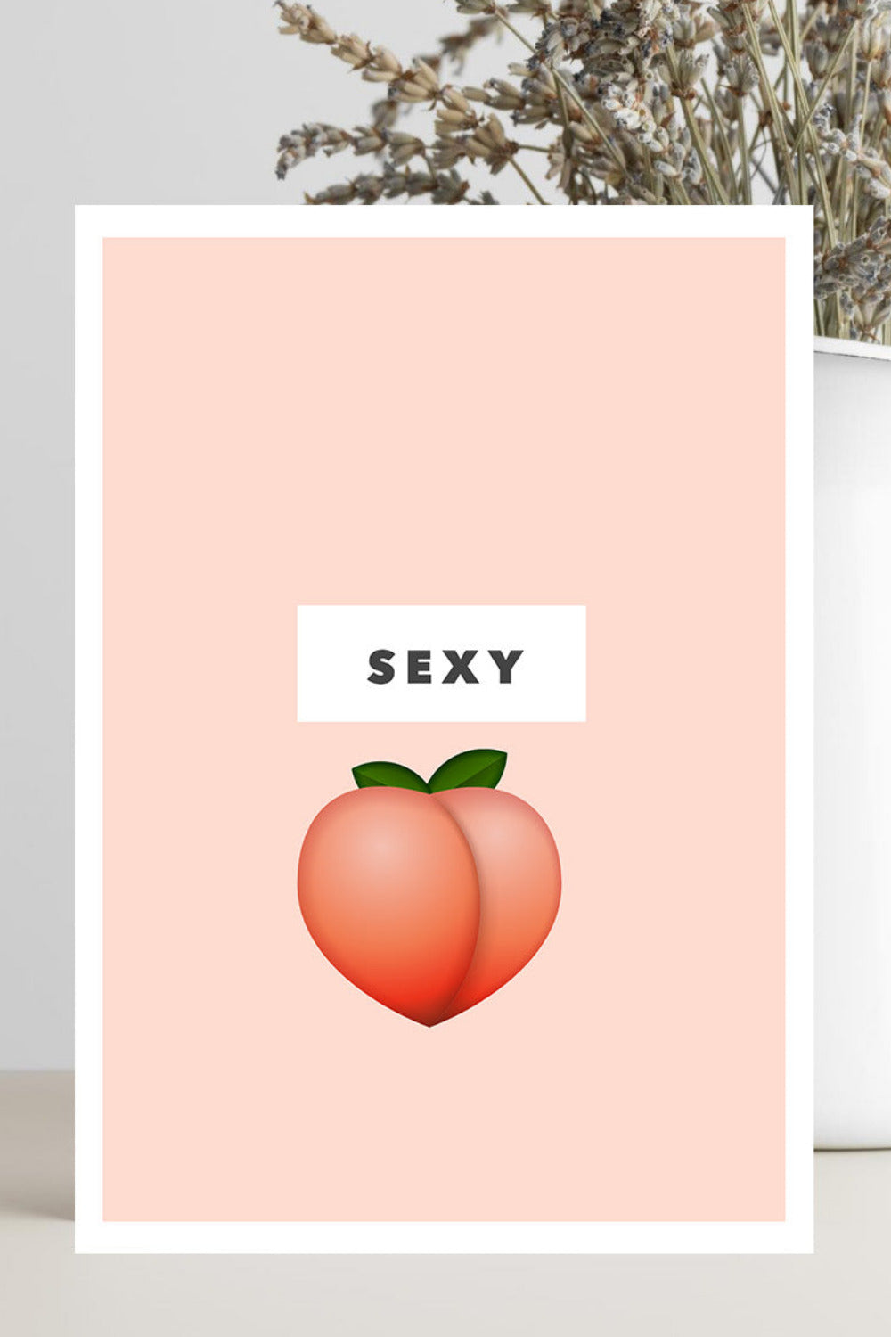 A vertical art print with a peach-colored background, featuring a white rectangular label with the word 'SEXY' above a peach illustration, displayed in a styled setting next to a white vase with dried flowers.