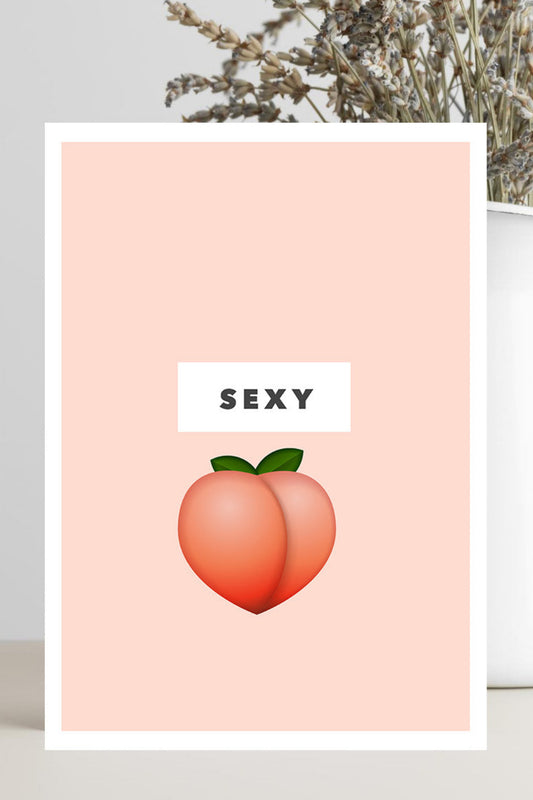 A vertical art print with a peach-colored background, featuring a white rectangular label with the word 'SEXY' above a peach illustration, displayed in a styled setting next to a white vase with dried flowers.