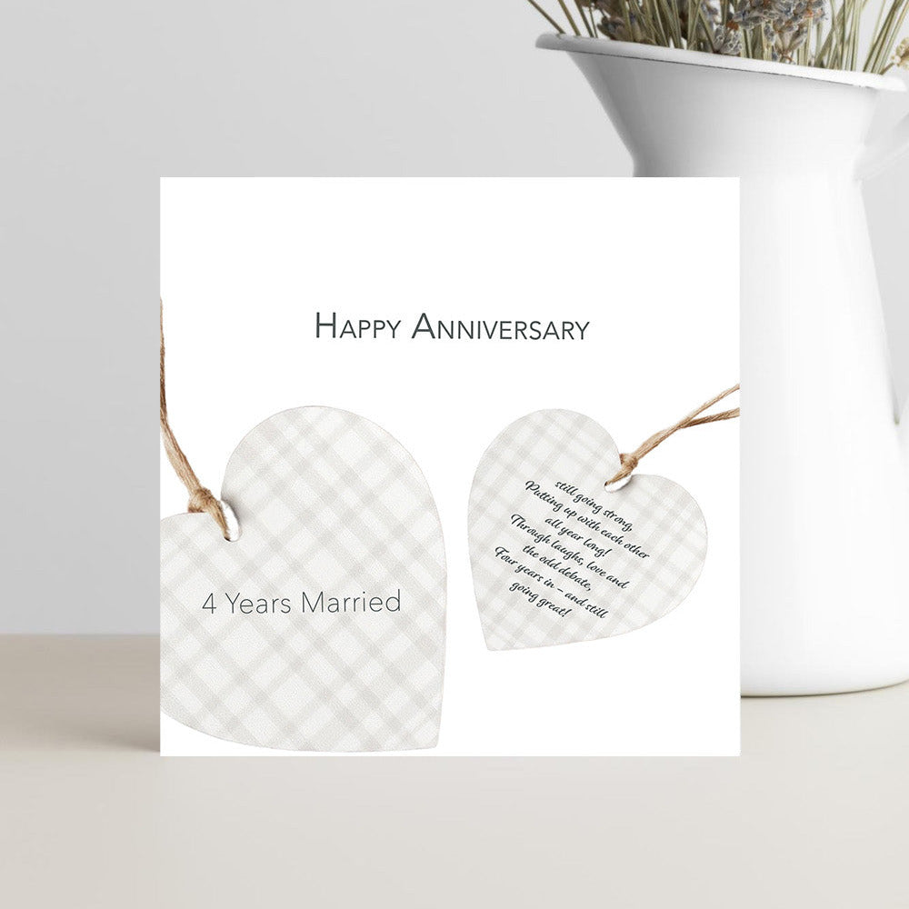 A square white anniversary card with the text 'Happy Anniversary' and '4 Years Married' above two heart-shaped hanging tags with a gingham pattern, styled in front of a white vase with dried flowers on a light surface.