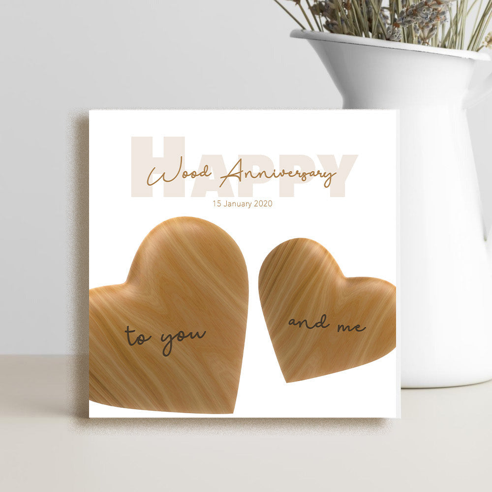 A square white greeting card with two wooden-texture hearts and the words 'HAPPY Wood Anniversary' and 'to you and me', styled in front of a white vase with dried flowers on a light gray surface.