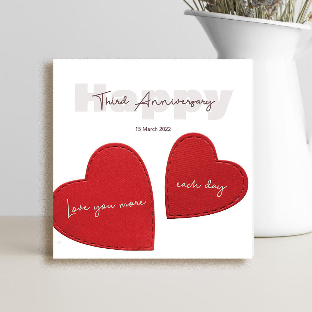 A square white anniversary card with two red heart shapes and cursive text reading 'Third Anniversary' and 'Love you more each day,' styled on a light surface beside a white vase with dried flowers.