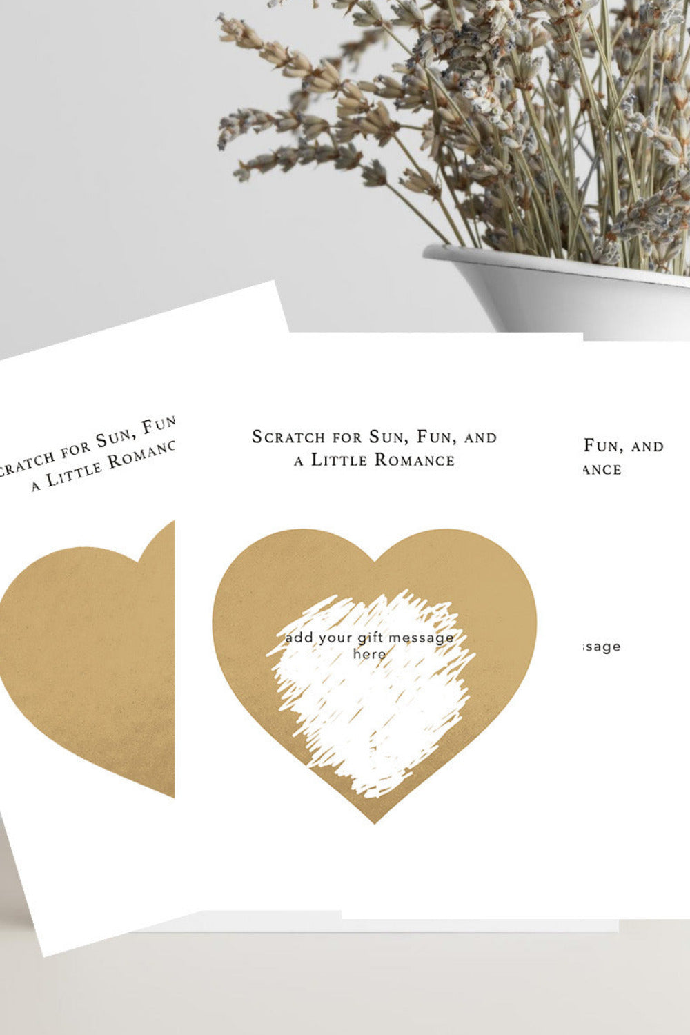 A white rectangular card with a gold heart-shaped scratch area and printed text, styled on a light surface with dried flowers in a white pot in the background.