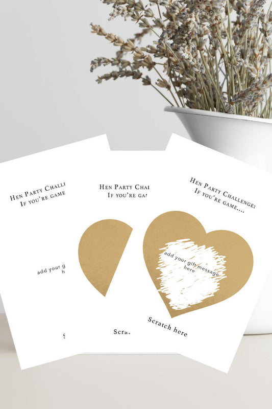 white party card with gold heart graphics and printed challenge text, styled on a light surface with dried flowers in a white vase in the background.