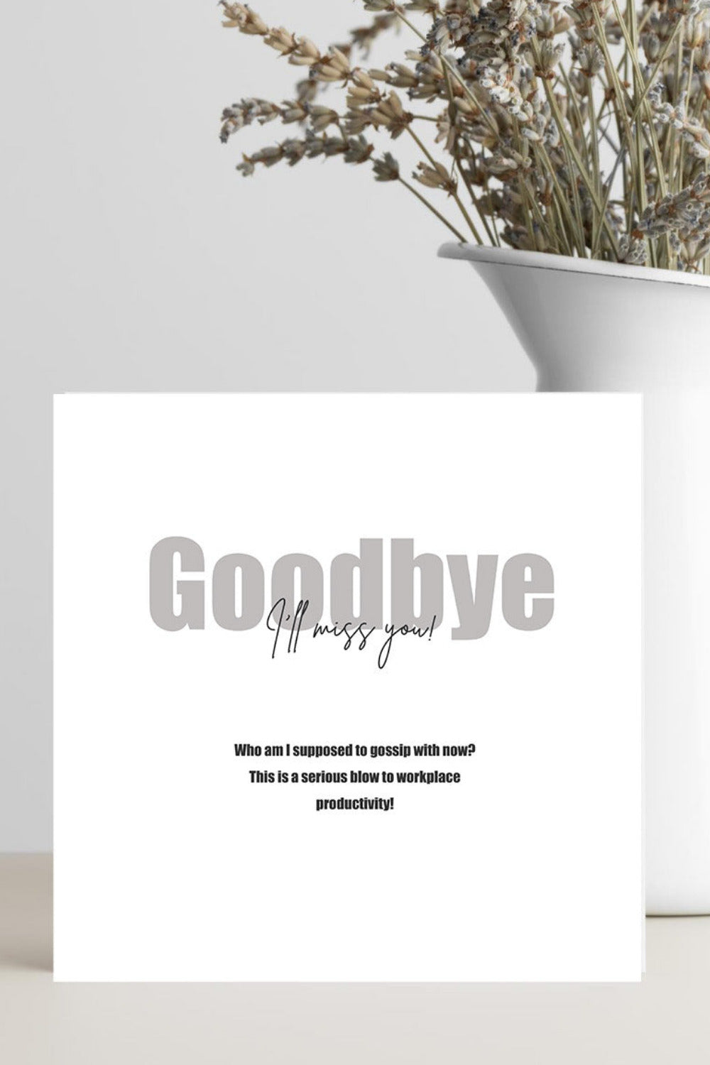 A white greeting card with the text 'Goodbye' and smaller farewell message, standing on a light surface beside a white vase filled with dried flowers against a light gray background.