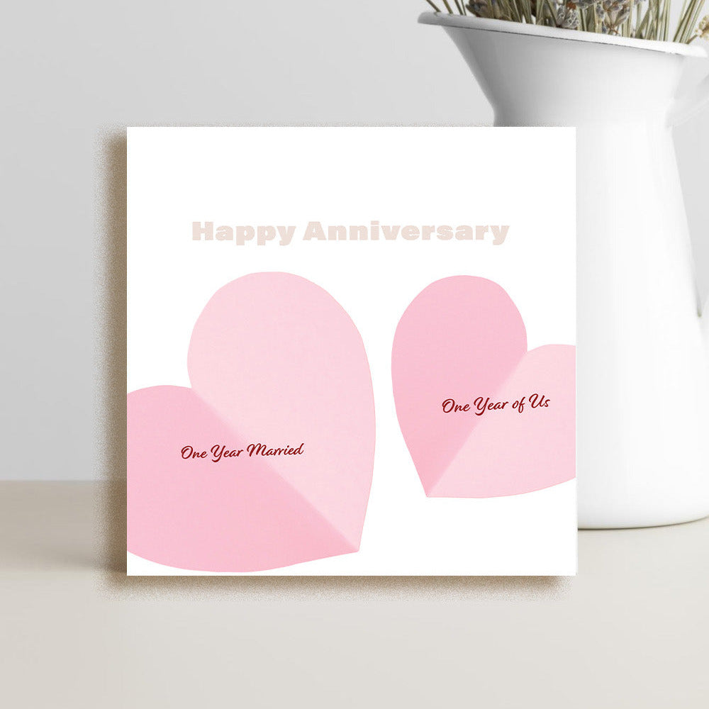 A square white greeting card with two pink heart shapes and the words 'Happy Anniversary' and 'One Year Married' / 'One Year of Us' stands on a light surface beside a white vase with dried flowers.