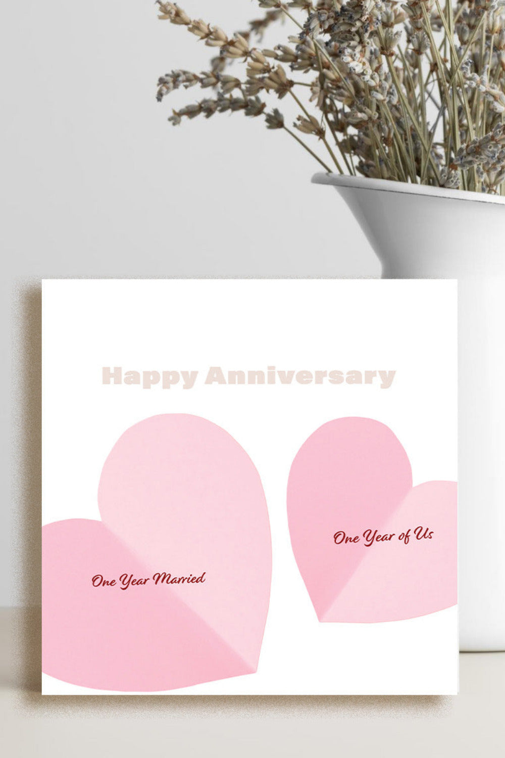 A square white greeting card with two pink heart shapes and the words 'Happy Anniversary' and 'One Year Married' / 'One Year of Us' stands on a light surface beside a white vase with dried flowers.