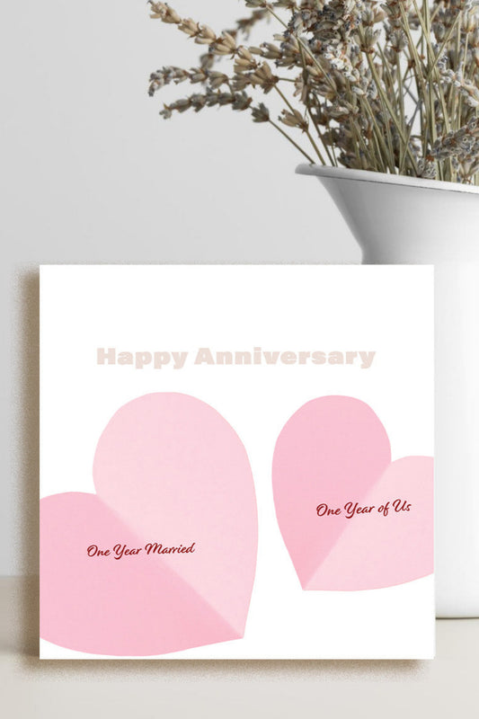 A square white greeting card with two pink heart shapes and the words 'Happy Anniversary' and 'One Year Married' / 'One Year of Us' stands on a light surface beside a white vase with dried flowers.