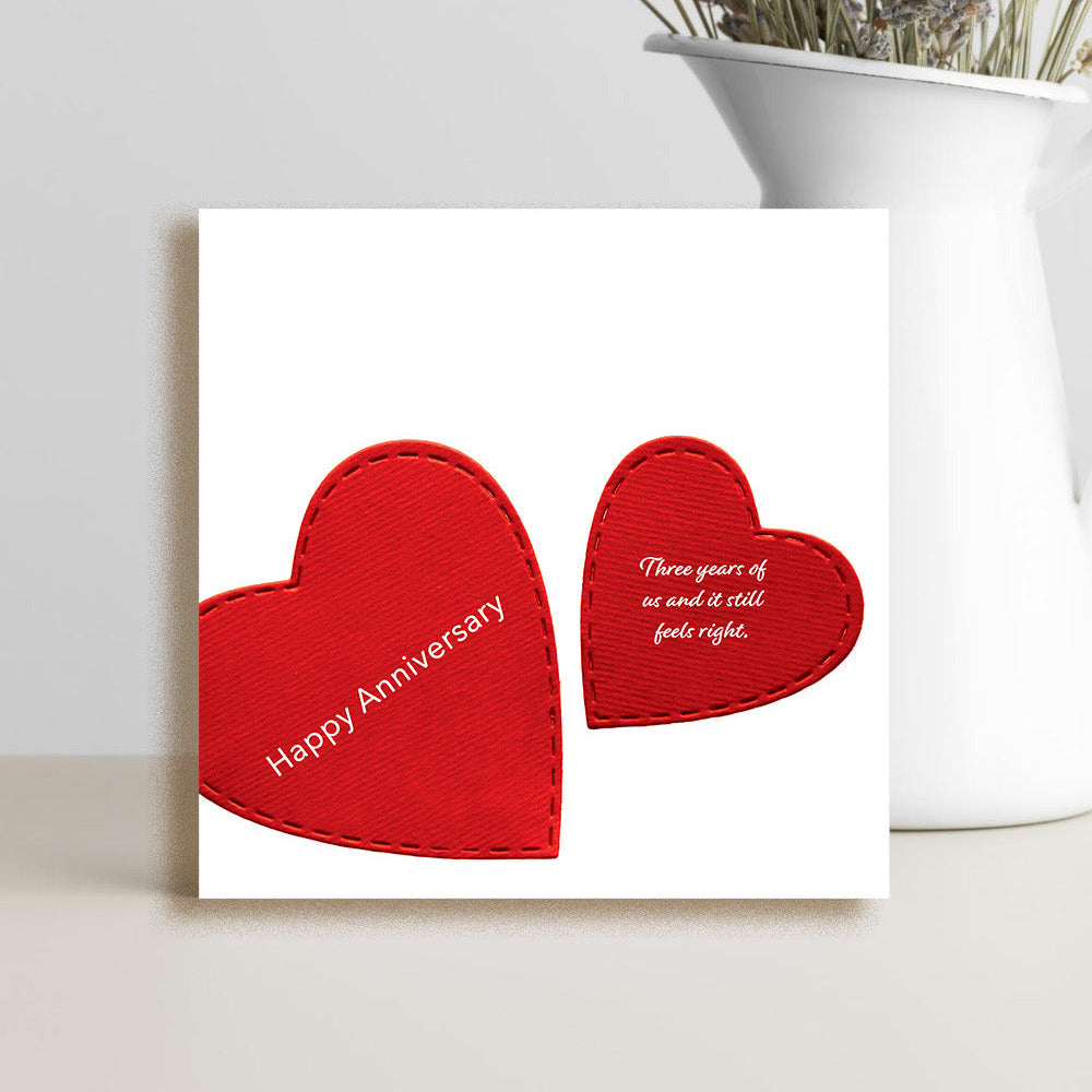 A square white greeting card with two red heart shapes and printed anniversary text, styled in front of a white vase with dried flowers on a light surface.