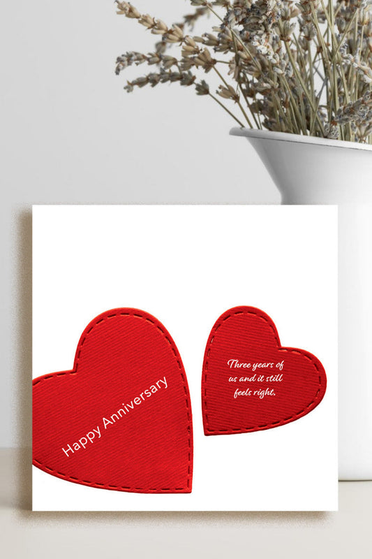 A square white greeting card with two red heart shapes and printed anniversary text, styled in front of a white vase with dried flowers on a light surface.