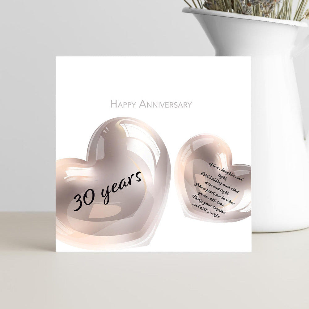 A square anniversary card with two glossy rose-gold heart shapes and the words 'Happy Anniversary' and '30 years,' displayed in front of a white vase with dried flowers on a light surface.