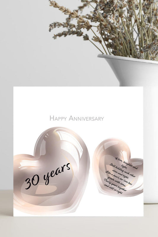 A square anniversary card with two glossy rose-gold heart shapes and the words 'Happy Anniversary' and '30 years,' displayed in front of a white vase with dried flowers on a light surface.