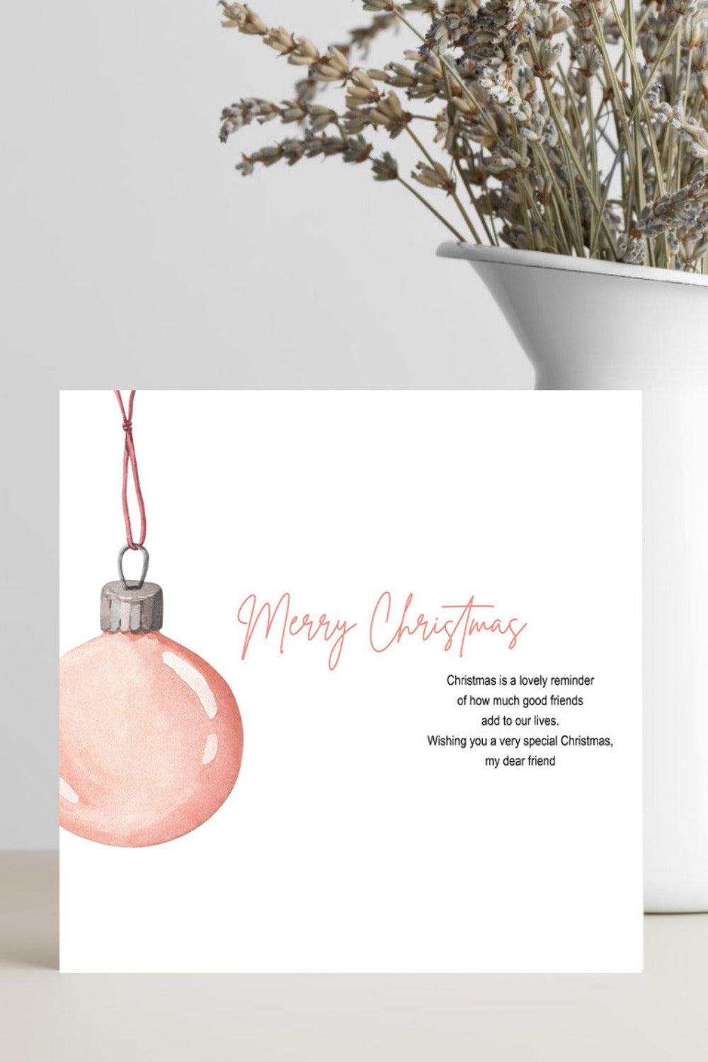 A square white Christmas greeting card with a watercolor pink ornament illustration and the words 'Merry Christmas' in pink script, styled on a light surface beside a white vase with dried flowers.