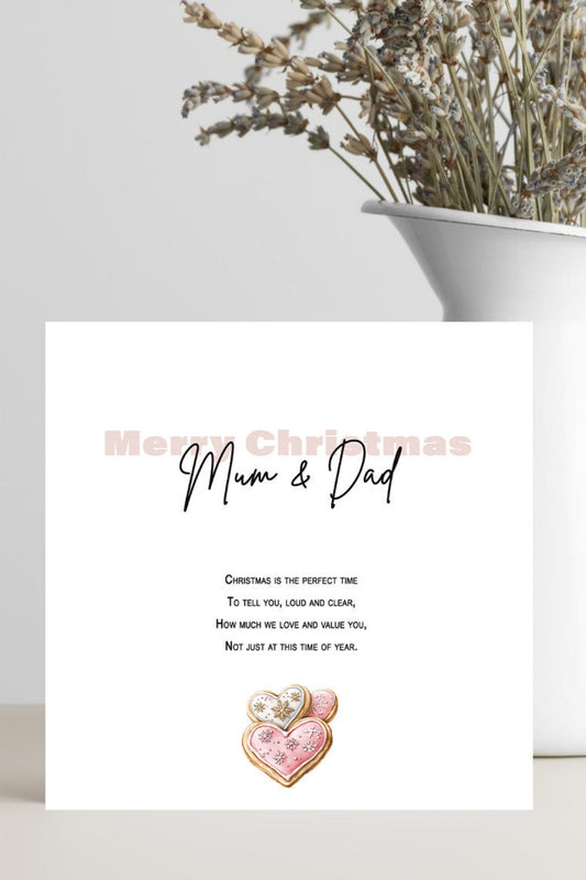 A white Christmas greeting card addressed 'Mum & Dad' with a small pink heart illustration, styled on a tabletop next to a white vase with dried flowers.