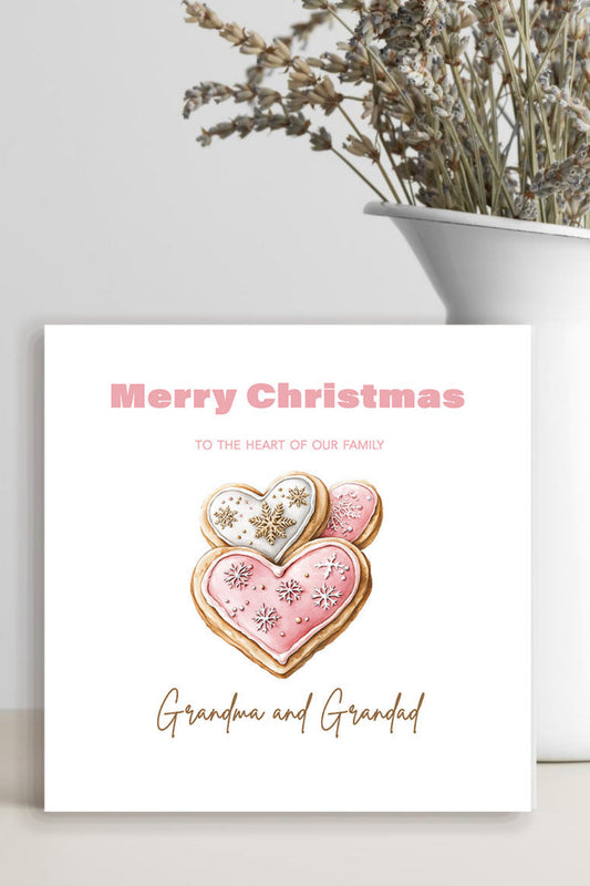 A square Christmas greeting card with a watercolor-style pink heart cookie illustration and the text 'Merry Christmas to the heart of our family' and 'Grandma and Grandad,' displayed on a light surface beside a white vase with dried flowers.