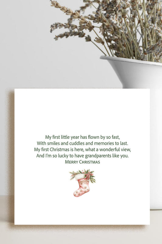 A square white greeting card with a sentimental Christmas message and a small red stocking illustration, styled on a tabletop next to a white vase with dried flowers.