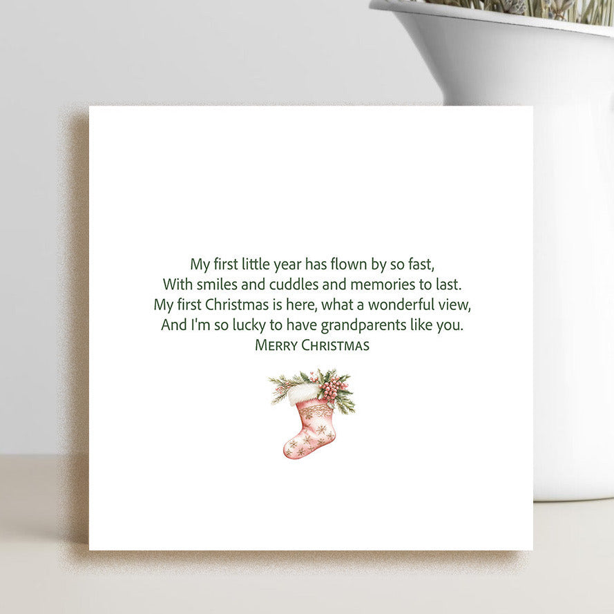 A square white greeting card with a sentimental Christmas message and a small red stocking illustration, styled on a tabletop next to a white vase with dried flowers.