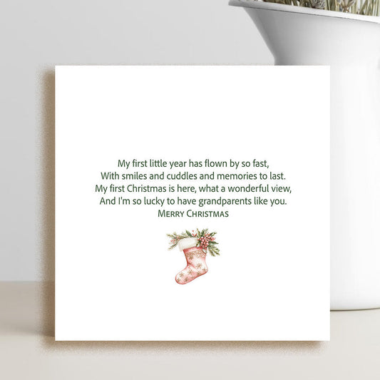 A square white greeting card with a sentimental Christmas message and a small red stocking illustration, styled on a tabletop next to a white vase with dried flowers.