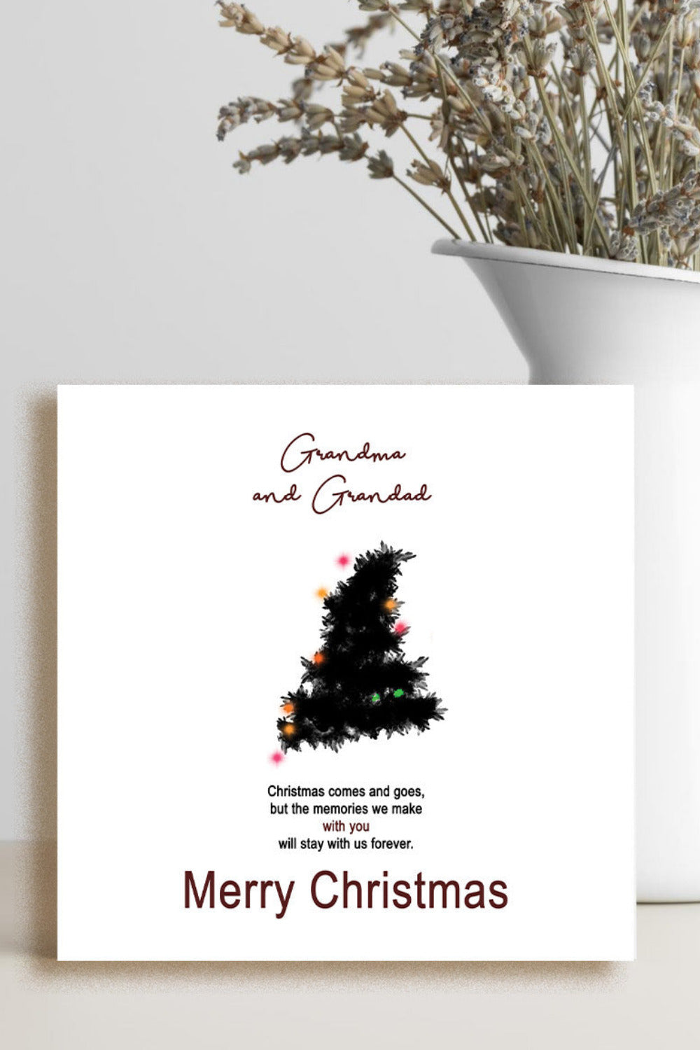 A personalized Christmas card with a black Christmas tree illustration and the text 'Grandma and Grandad' and 'Merry Christmas', displayed on a light surface next to a white vase with dried flowers.