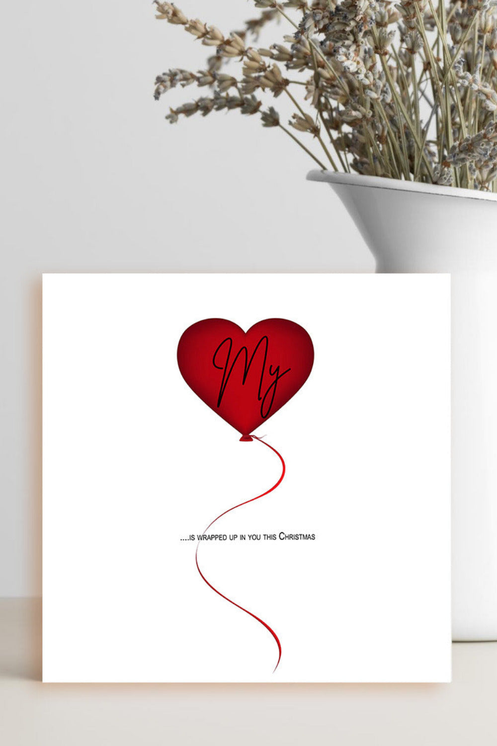 A square white art print or card featuring a red heart-shaped balloon with the word 'My' in black script and a red string, displayed on a tabletop next to a white vase with dried flowers.