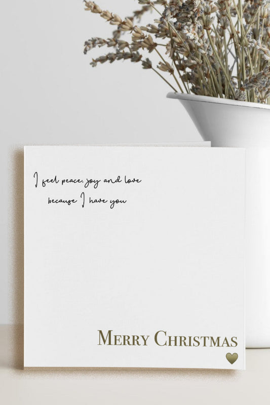 A square white Christmas greeting card with elegant black script text reading 'I feel peace joy and love because I love you' and 'Merry Christmas' in gold at the bottom, styled on a light surface next to a white vase with dried flowers.