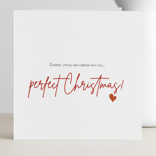 A white greeting card with the text 'perfect Christmas!' in red script and a small red heart, standing on a light surface beside a white vase with dried flowers.