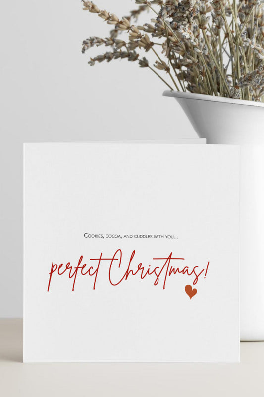 A white greeting card with the text 'perfect Christmas!' in red script and a small red heart, standing on a light surface beside a white vase with dried flowers.