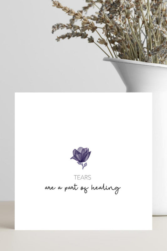 Card with a purple flower illustration and text 'Tears are a part of healing' on a white surface with a vase of dried flowers in the background.