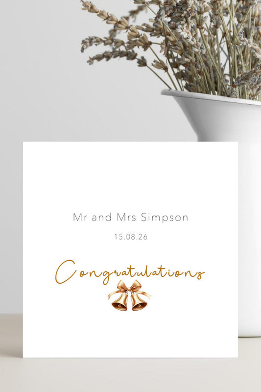 A personalized white wedding congratulations card with gold script text and a small gold bell illustration, styled on a light surface next to a white vase with dried flowers.