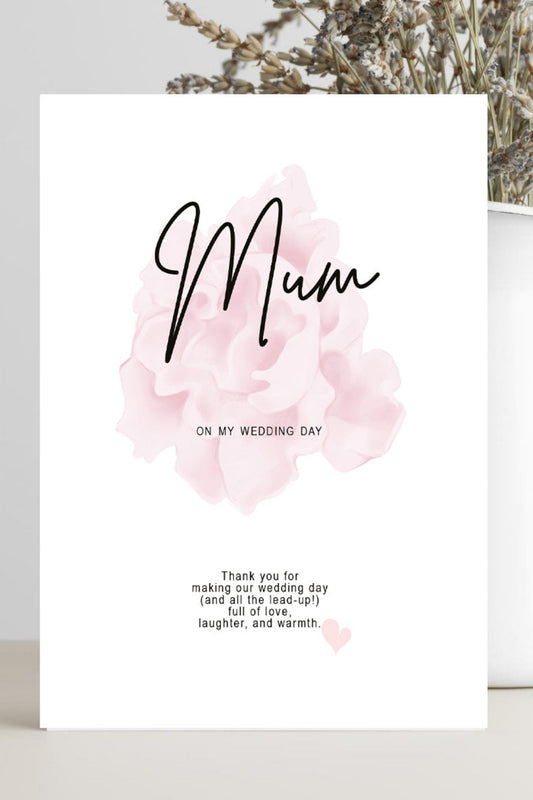 A white greeting card with a pink watercolor flower and the word 'Mum' in black script, standing on a light surface with dried flowers in a white vase behind it.