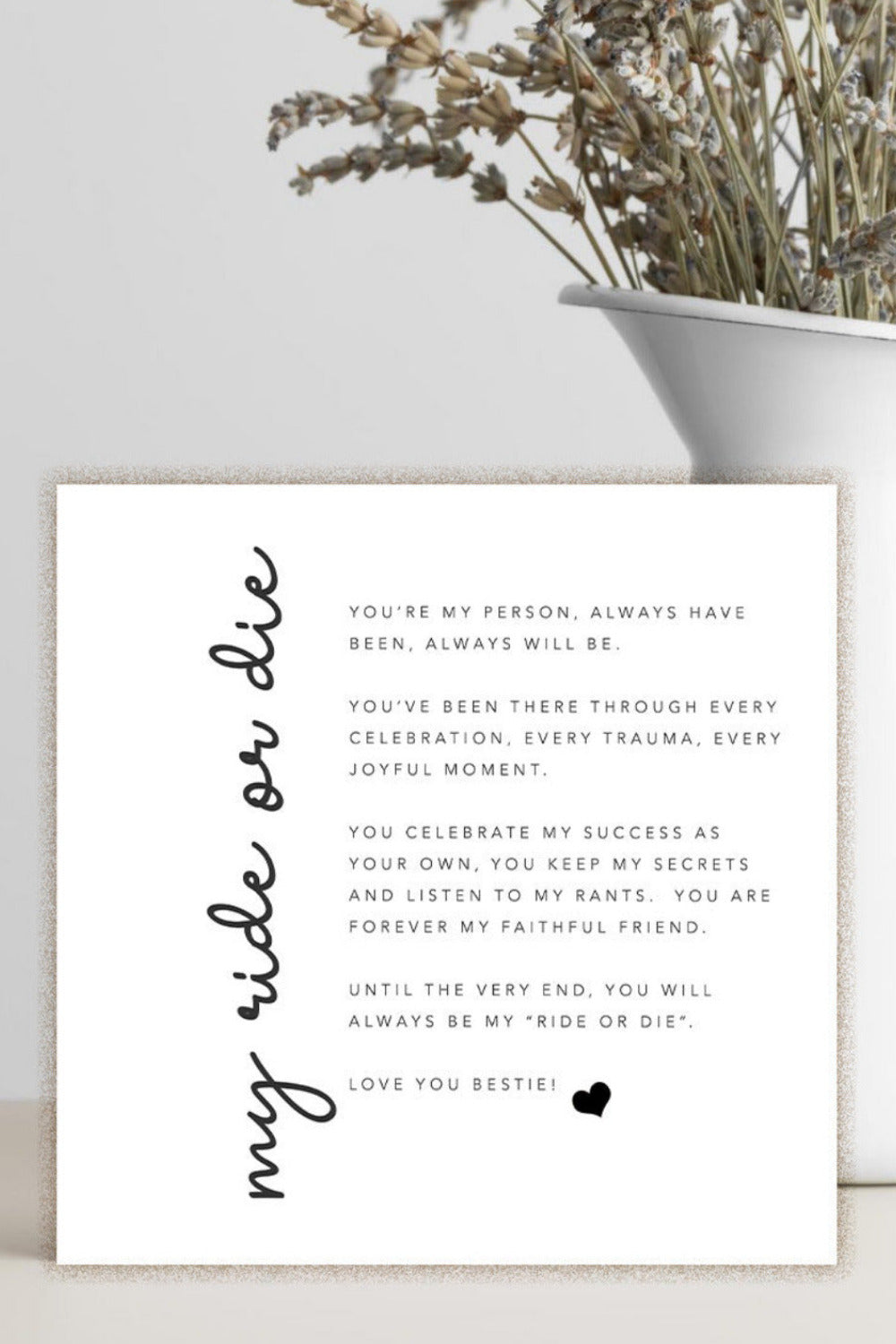 A white square card with black printed text and the phrase 'may ride or die' in script, styled on a light surface beside a white vase with dried flowers.