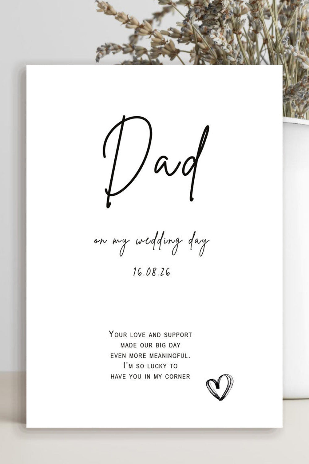 A white wedding guest book card with black script text reading 'Dad on my wedding day' and a small heart at the bottom, styled in a vase with dried flowers on a light gray surface.