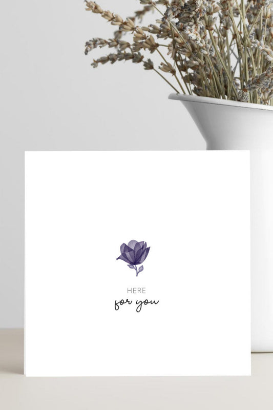 Card with a purple flower illustration and 'Here for you' text, placed on a surface with a vase of dried flowers in the background.