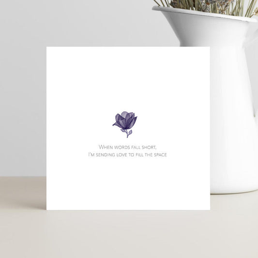Greeting card with a purple flower illustration and text on a white surface with a vase of dried flowers in the background.