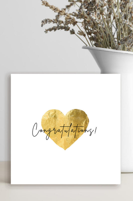 A square white congratulations card with a textured gold foil heart and the word 'Congratulations!' in black script, styled on a light surface next to a white vase with dried flowers.