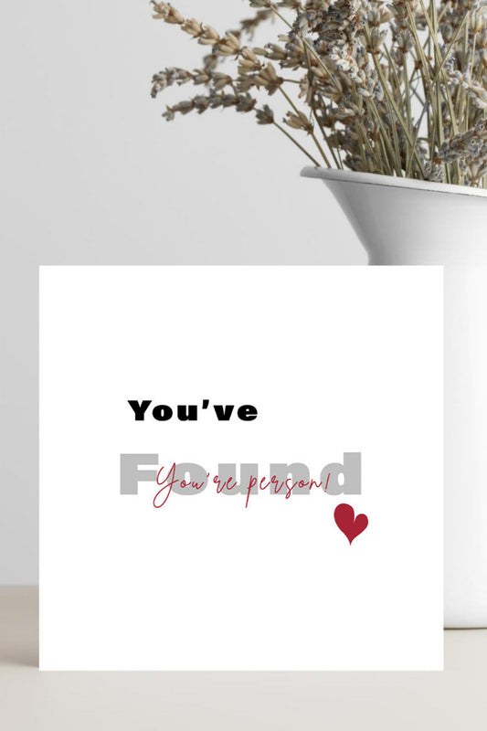 A white greeting card with the text 'You've found You're person!' in black and pink script, with a small red heart, styled on a light surface next to a white vase with dried flowers.