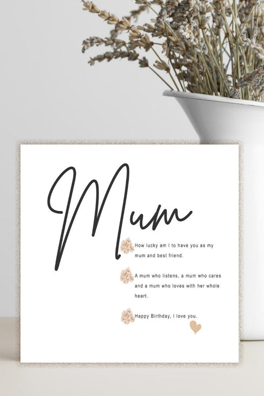 A square white greeting card with the word 'Mum' in large black script and a short sentimental message below, styled on a tabletop beside a white vase with dried flowers against a light gray wall.