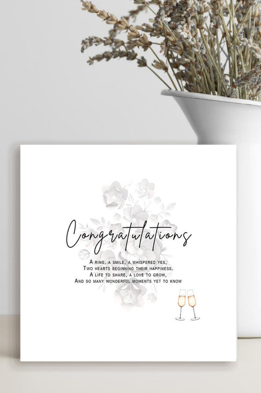 A square white congratulations card with elegant black script and small floral accents, leaning against a light wall beside a white vase filled with dried flowers.