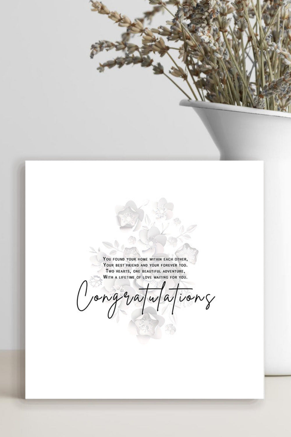 A square white congratulations card with a subtle floral design and cursive text, styled on a light beige surface next to a white ceramic vase containing dried flowers against a light gray wall.