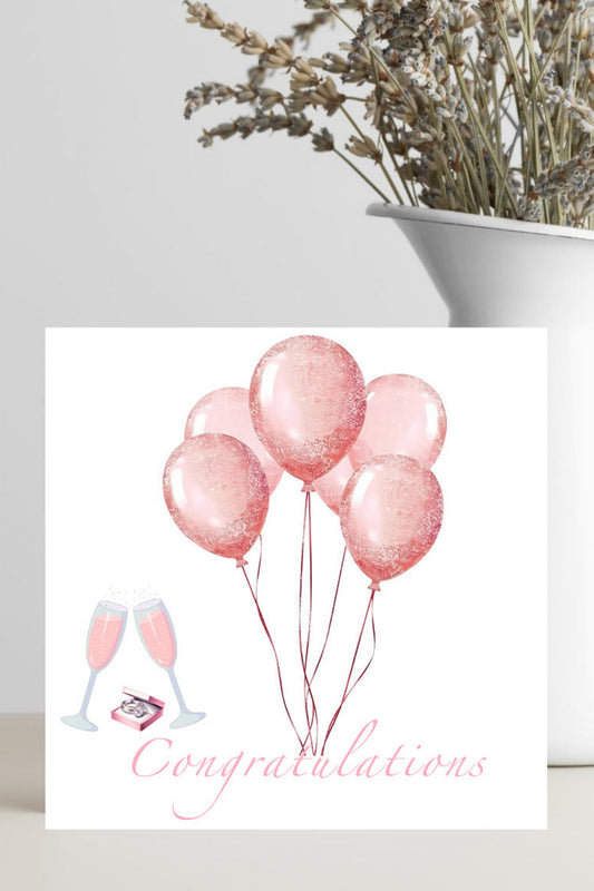 Greeting card with pink balloons and 'Congratulations' text on a white surface with a plant in the background.
